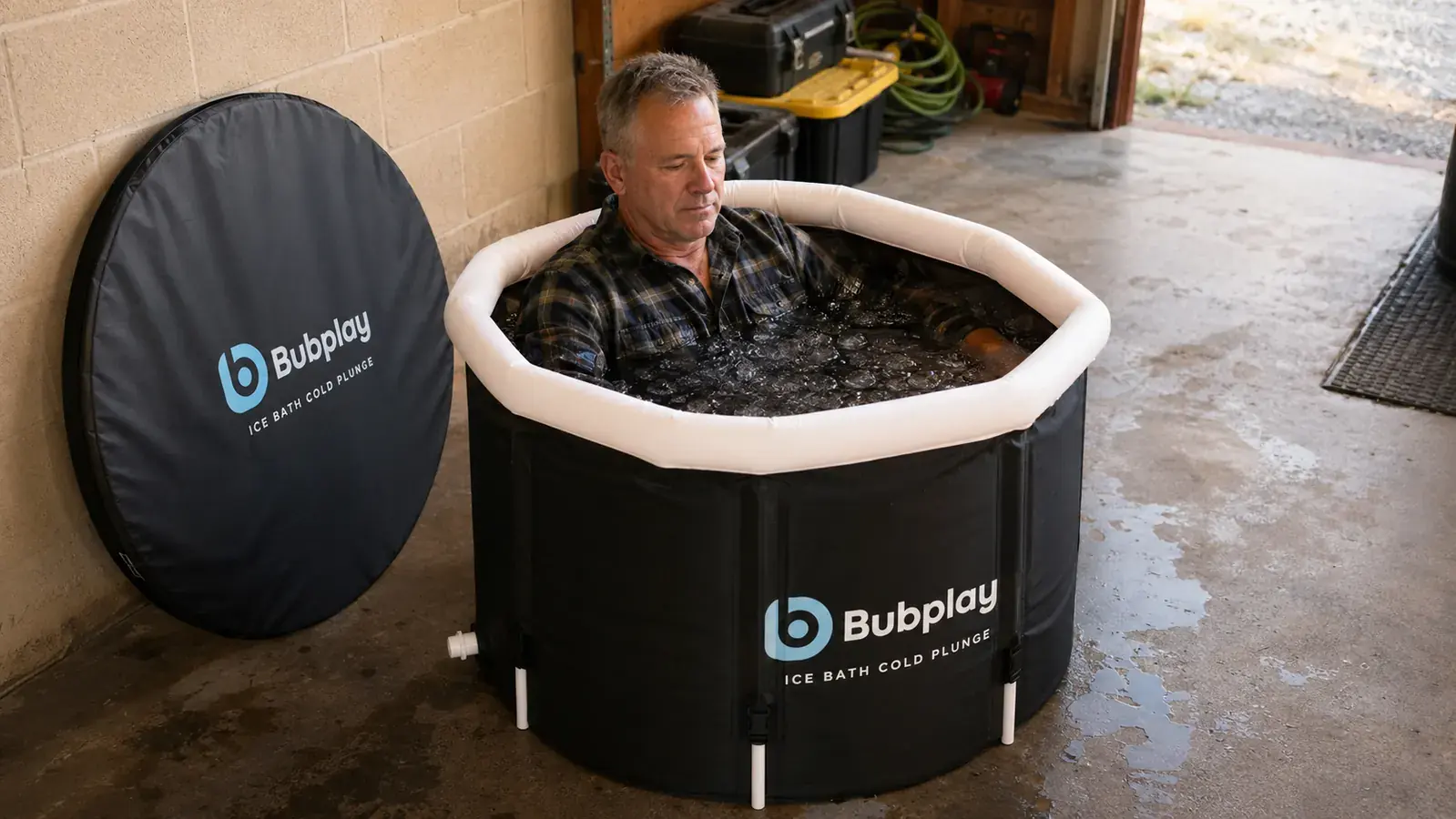 Man in work boots and joggers sitting chest-deep in the Bubplay cold plunge tub with a timer on the tub cover beside him